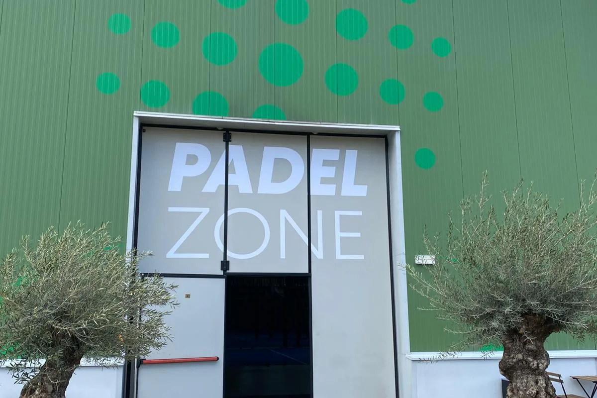 Intrarea în Padel Zone Oradea: ușă albă cu sigla clubului, pe perete verde, între doi măslini